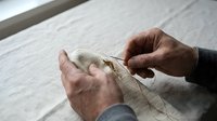 Senior craftsman hands carefully threading needle through natural linen fabric, showcasing traditional textile work and artisan skills in bright workshop setting.