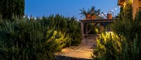 Wooden boardwalk path through coastal dunes with native vegetation leading to beach house at twilight with warm lighting and clear blue sky.