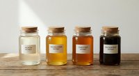 Four glass jars with cork lids containing different honey varieties from light to dark amber, labeled and arranged on wooden surface for gourmet food display.