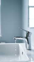 Chrome bathroom faucet with running water over white sink basin, bright natural light from window, modern minimalist interior design for home improvement and plumbing concepts.