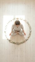 Woman in beige linen clothing positioned within circular arrangement of seashells and natural elements on light wooden floor, overhead view for wellness and mindfulness concepts.