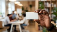 Male professional with dark skin tone holding blank white business card in modern office environment with colleagues working at desks in background for corporate branding and networking purposes.