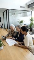 Three mature women professionals reviewing documents at wooden table in modern office with glass partitions and plants.