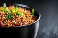 Spicy Asian noodles with minced meat sauce garnished with fresh herbs in black ceramic bowl against dark background, selective focus on steaming dish.