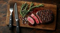 Grilled medium rare beef steak sliced on wooden cutting board with rosemary sprigs, black pepper, and stainless steel cutlery against dark rustic background.