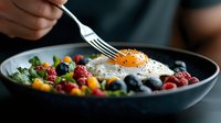 Healthy breakfast bowl with sunny side up egg, fresh berries, blueberries, raspberries and green salad mix served in dark ceramic plate. Fork reaching for food.