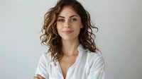 Young woman with wavy brown hair and Mediterranean ethnicity wearing white button-up shirt, smiling at camera against neutral background. Suitable for business, lifestyle, and professional portraits.