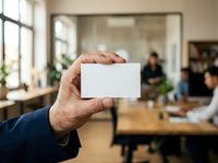Man holding blank white business card in modern office environment with colleagues working in background.