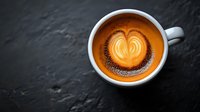Heart-shaped latte art in white ceramic cup on dark stone background, golden coffee crema with delicate milk foam pattern and chocolate powder garnish.