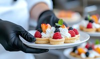 Pastry chef in black gloves holding white plate with gourmet tarts topped with fresh raspberries, blackberries, whipped cream and fruit garnish in professional kitchen setting.