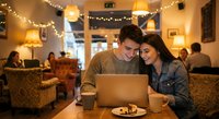 Young couple sharing laptop computer in cozy cafe with warm lighting and string lights creating romantic atmosphere for date or work session.