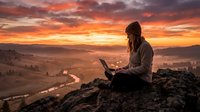Woman with long dark hair, wearing casual clothing, sitting on a rocky mountain outcrop while working on laptop at sunset. Golden hour landscape with winding river and misty valley below. Remote work, freelance, and digital nomad concept.