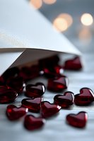 Red glass hearts scattered from white paper curl against blurred bokeh background, creating romantic Valentine's Day mood with soft lighting and shadows.
