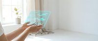 Person holding transparent digital tablet with holographic interface displaying data visualization and network connections in bright, modern workspace with natural light from window.