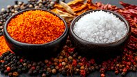 Orange and white spices in black bowls surrounded by scattered peppercorns, dried chili peppers and aromatic seasonings on dark background, macro food photography.