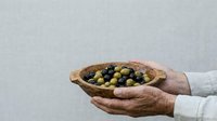 Hands holding wooden bowl filled with mixed green and black olives against neutral background, isolated for design use.