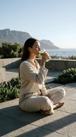Woman with long dark hair, appearing to be of Mediterranean or South Asian descent, sitting cross-legged on a terrace drinking water. Scenic coastal landscape with mountains visible behind her. Wellness and hydration concept for lifestyle and health content.
