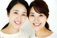 Two young Asian women with bright genuine smiles wearing white tops against light background, showcasing natural beauty and friendship in casual portrait.