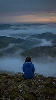 Woman in blue jacket sitting on rocky mountain peak overlooking misty forested valleys at dawn, contemplating vast landscape with fog-covered hills and dramatic sky.