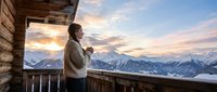 Woman in warm sweater holding child on mountain cabin balcony at sunset, snow-covered peaks in background, family travel and alpine lifestyle concept.