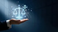 Businessman hand holding glowing scales of justice symbol with particles and light effects on dark blue background for legal services and law concepts.