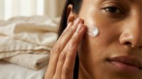 Young woman applying white moisturizing cream to face during morning skincare routine in bright bedroom. Natural beauty and self-care concept.