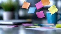 Colorful sticky notes floating in air against blurred office background with laptop and coffee cup, creating dynamic composition for business planning.