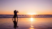 Romantic couple dancing on beach at sunset with golden sky and ocean waves reflecting on wet sand creating magical silhouette moment for love and romance themes.
