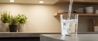 Young woman pouring fresh sparkling water into glass on modern kitchen counter with potted plants and shelving in background, hydration and healthy lifestyle concept.