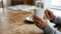 Senior woman with brown skin holding white mug at wooden table, wearing gray sweater sleeves, daytime indoor setting for lifestyle, wellness, and beverage marketing.