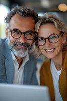 Mature couple with glasses smiling while looking at laptop, representing successful business partnership or retirement planning.