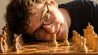 Young Caucasian man with curly hair and glasses lying next to wooden chess set, contemplating strategic moves in intellectual board game competition.
