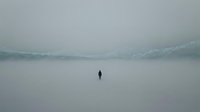 Solitary figure standing on frozen lake surrounded by misty mountains and fog, conveying isolation and contemplation in winter landscape.