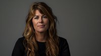 Professional woman with long wavy brown hair wearing black top against dark gray background in dramatic studio lighting, looking confident and approachable in corporate headshot style.
