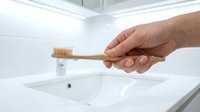 Hand holding eco-friendly bamboo toothbrush over white bathroom sink. Sustainable oral hygiene and zero waste lifestyle concept for dental care routine.