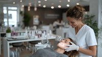 Professional beautician performing eyebrow shaping treatment on female client in modern beauty salon with soft lighting and elegant interior design.