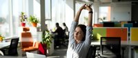 Young Caucasian businesswoman stretching arms above head at office desk with laptop, taking break from work in modern open space.
