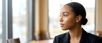 Young African American businesswoman in black blazer looking thoughtfully out office window during daytime, professional corporate environment.