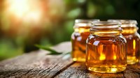 Glass jars filled with golden honey on rustic wooden surface against blurred natural background, sunlight creating warm amber glow through transparent containers.