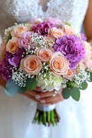 Bridal bouquet with peach roses, purple hydrangeas, and delicate baby's breath against white wedding dress background, held by bride's hands in soft focus.