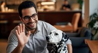 Young man with dark hair and glasses wearing light gray shirt waving while posing with dalmatian dog indoors, suitable for pet care, veterinary services, animal companionship and lifestyle content.