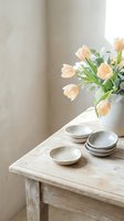 Soft peach tulips in ceramic vase with handmade pottery bowls on rustic wooden table creating serene minimalist home decor scene with natural lighting.