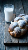Fresh braided challah bread dusted with powdered sugar served with glass of milk on rustic wooden table for traditional breakfast or bakery display.