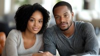 Young African American couple smiling at camera, wearing casual sweaters while relaxing at home. Natural lighting creates warm, authentic atmosphere for lifestyle content.
