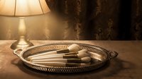 Luxury makeup brushes with white bristles arranged on a vintage silver tray, warm lamp glow, elegant vanity table setting.