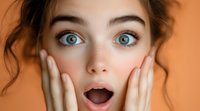 Young Caucasian woman with bright blue eyes and natural makeup expressing surprise with open mouth and hands on cheeks against orange background.