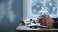 Senior man hands protecting glass dome cloche on wooden table near window with natural light for insurance protection security concept.