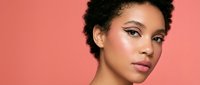 Young mixed-race woman with curly hair and natural makeup looking at camera against coral pink background for beauty and skincare campaigns.