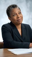 Confident African American businesswoman in dark suit sitting at desk with documents, looking up thoughtfully in modern office environment.