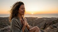 Peaceful woman with curly hair meditating at sunrise on rocky coastline, eyes closed in serene contemplation during golden hour by ocean.